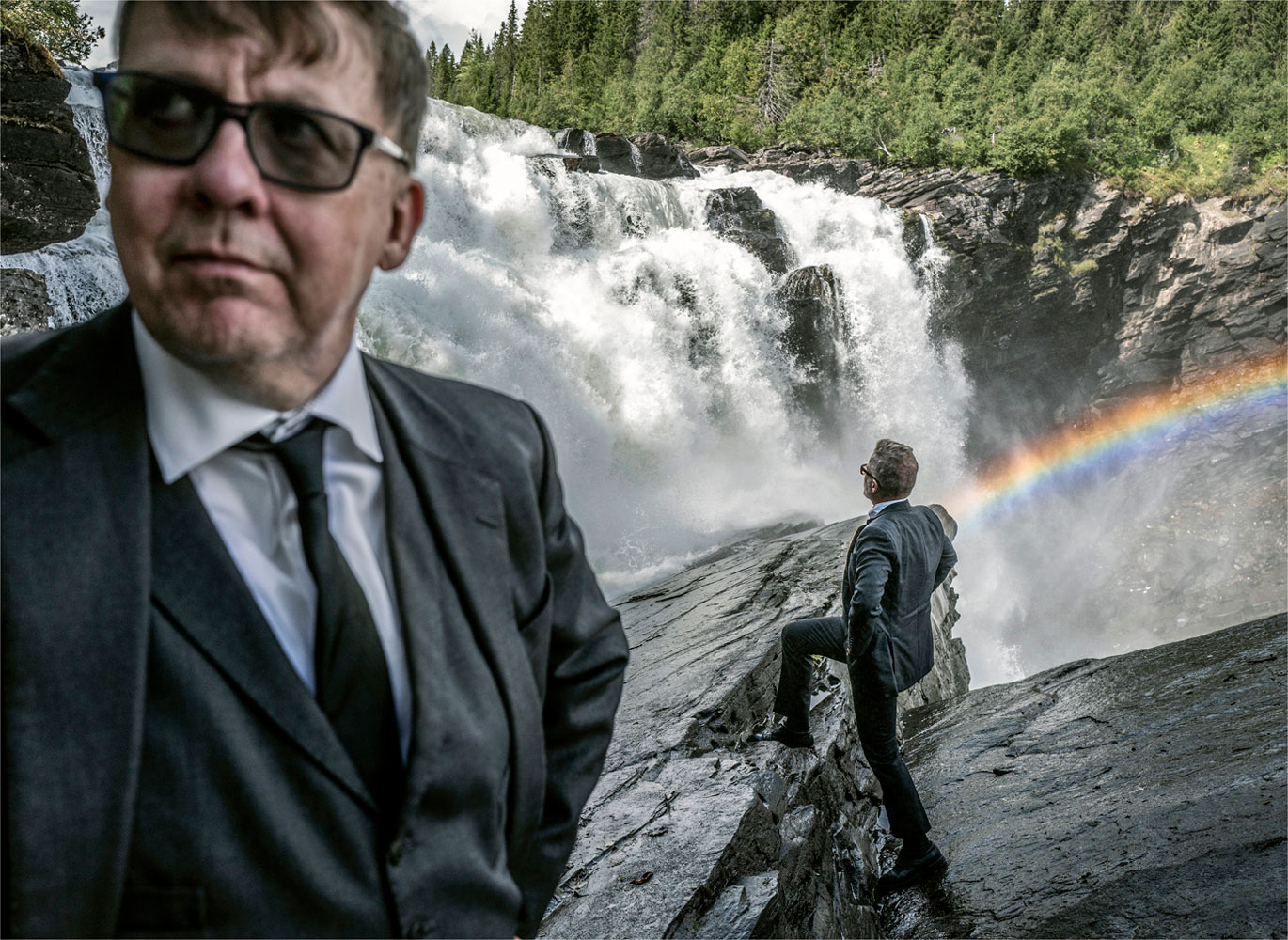 Foto. En kostymklädd man i halvfigur syns till vänster. Han har solglasögon och tittar snett utanför bild. Till höger bakom honom står en till kostymklädd man på en klippa. I bakgrunden syns ett mäktigt vattenfall. En regnbåge syns vid vattenfallet. 