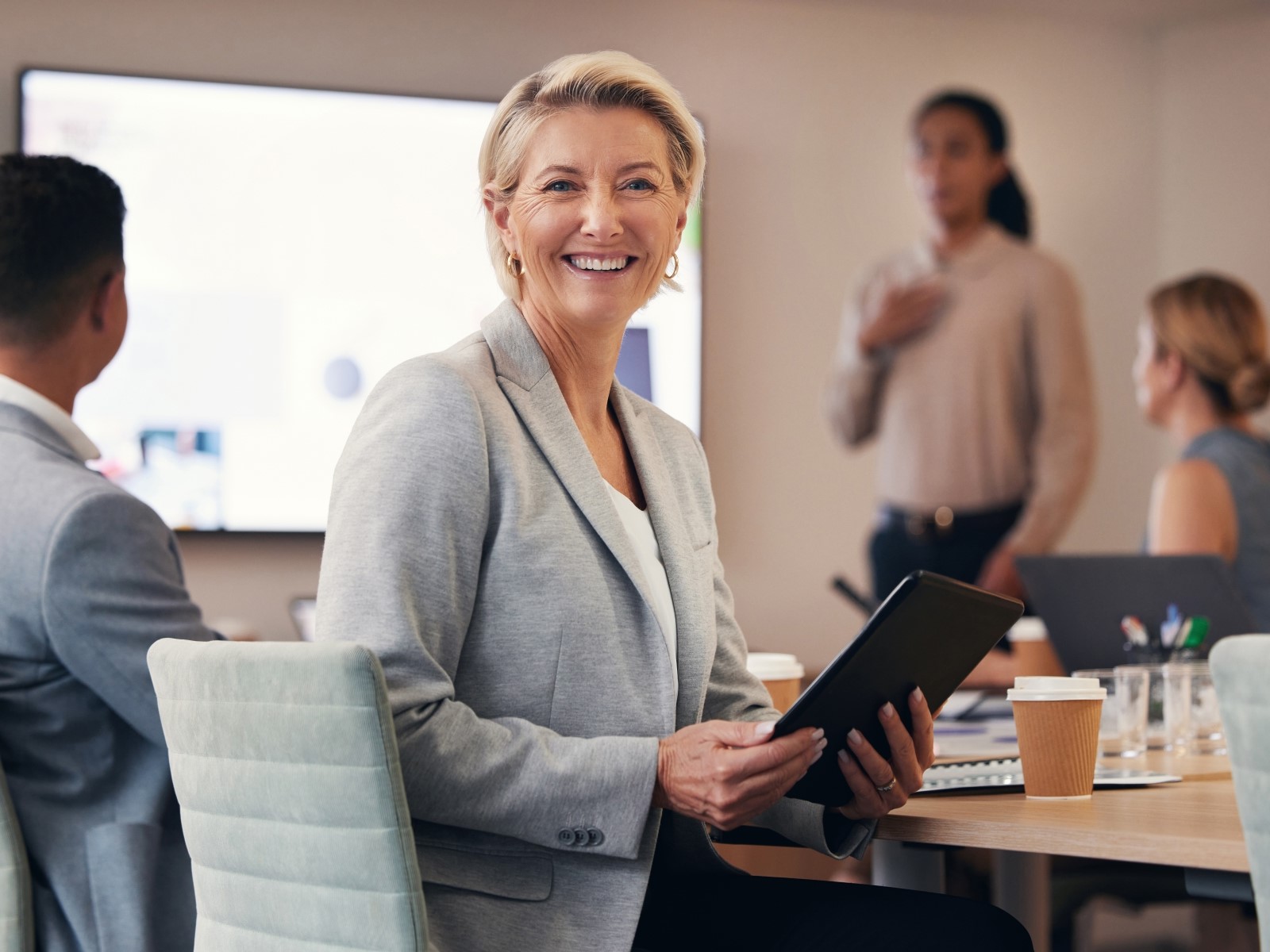Glad kvinnlig företagare tittar rakt mot kameran. Sitter i mötesrum där någon håller en presentation i bakgrunden. 
