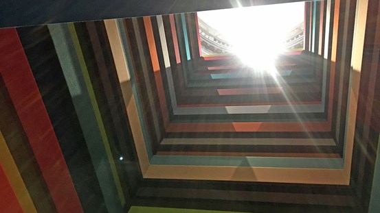 Ljus skiner ner mot betraktaren i en öppning längst upp i ett schakt med väggar med vertikala ränder i olika färger. 