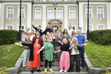 Ett gäng personal från Örnsköldsviks folkhögskola står på en trappa framför skolbyggnaden med utsträckta armar. 