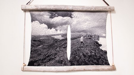 En textil i svartvitt är uppspänd mellan två trästammar upptill och nertill. Motivet visar ett vidsträckt landskap med en person som går till höger i bild. Allt i en grov pixelrastrering. I mitten av textilen sträcker sig ett avlångt vertikalt hål där den vita väggen syns.