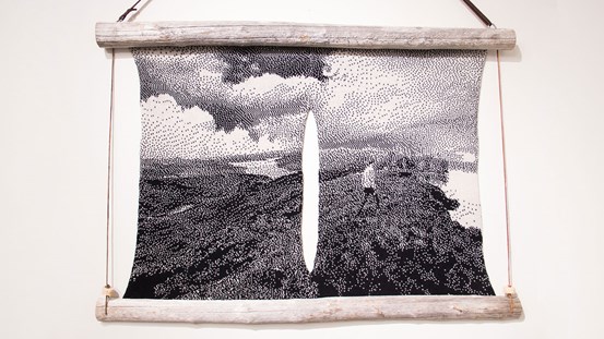 En textil i svartvitt är uppspänd mellan två trästammar upptill och nertill. Motivet visar ett vidsträckt landskap med en person som går till höger i bild. Allt i en grov pixelrastrering. I mitten av textilen sträcker sig ett avlångt vertikalt hål där den vita väggen syns.