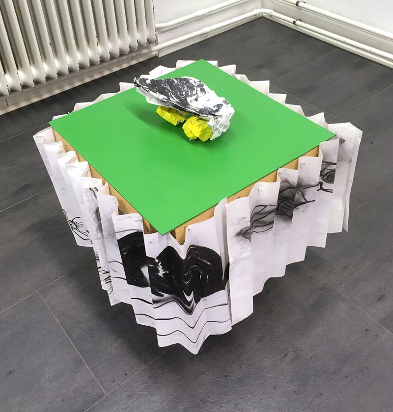 Foto. På ett grått golv står ett bord. Sidorna är täckt med veckat vitt papper eller textil som det ser ut att vara målat eller tryck i svart på. Bordskivan är grön och på den ett skulpturalt objekt i gult, vitt och svart. 