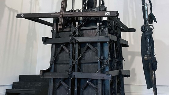 En stor maskinliknande träkonstruktion i svart med kedjor, vajrar, tyg och någon sorts metallkonstruktion. 