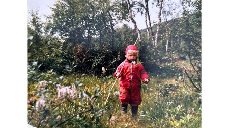 En liten flicka i röd overall och mössa i en skogsglänta. Hon bär en lång pinne över axeln. 