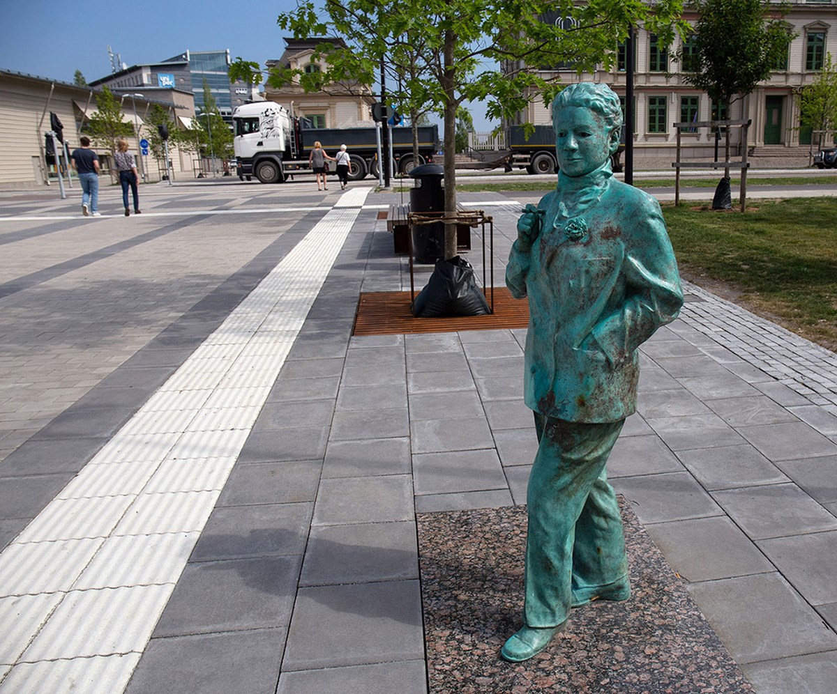 Foto av skulptur av Kai Gullmar. Skulpturen är ärgat grön ståendes  på trottoaren. Iförd herrkostym, halsduk och en blomma i slaget.  Hon har en cigarett i högerhanden och den vänstra nedstucken i  kavajfickan. 