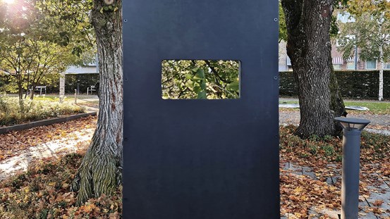 Park med träd, gångvägar och gulröda löv på marken. Mitt i bilden syns en del av periskopet. Det sträcker sig nerifrån och upp i bild, på det syns en rektangulär öppning, i den ser man de gröna trädkronorna.
