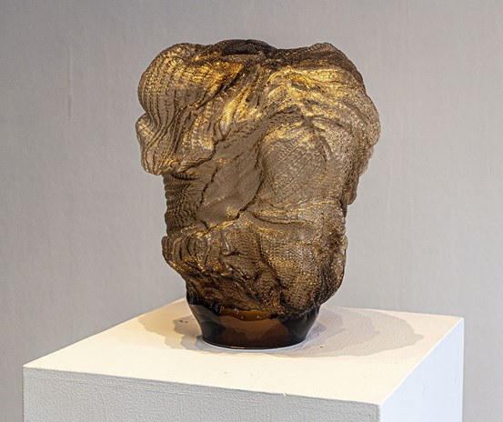 En bruntonad glasskulptur på ett podium. Formerna är böljande och organiska. Det räfflade ytmönstret liknar stickning eller virkning.