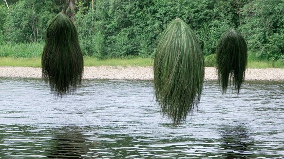Tre objekt, volymer av grönt långt gräs, hängandes ovan en vattenyta. I bakgrunden syns en stenig strand och skog. 