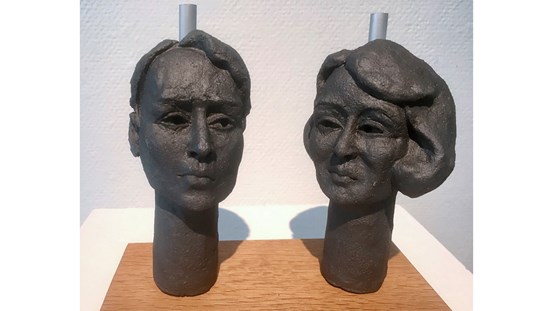 På en träplatta står två mörka keramikskulpturer. Det är två huvuden, ett manshuvud till vänster och ett kvinnohuvud till höger. Ovan bakom båda skymtar ett pinne, ett handtag, i ett grått material. I bakgrunden en vit vägg. 