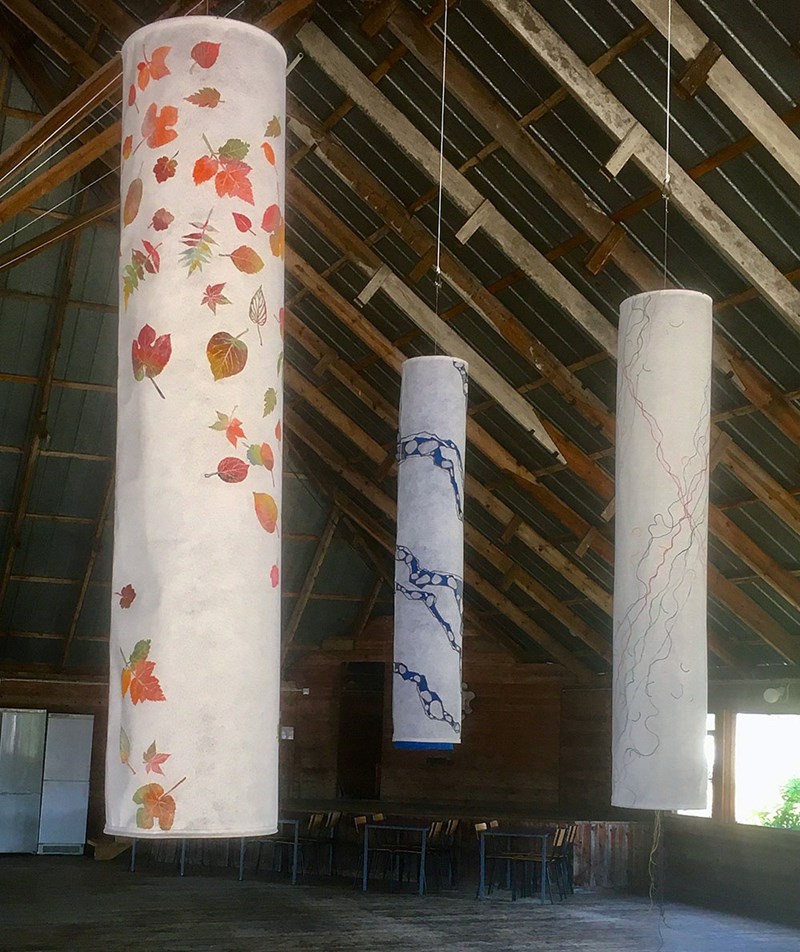Tre stycken cylinderformade verk hänger från taket, gjorda i ljus/beige fiberduk och med olika tryck på. Den längst fram till höger har tryck löv i olika färger, den längst bort i mitten har en lång form i blått som går runt verket. På den tredje till höger är det tryckt vad som ser ut som trådar i olika färger. 