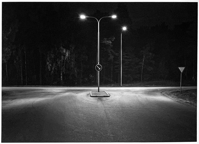 Ett svartvitt foto på kvällen. Vy mot en upplyst T-korsning. En väg löper mot betraktaren, en till höger och en till vänster. I mitten en liten refug med en hög stolpe som grenar sig i två längst upp med en gatlampa i vardera änden. På stolpen sitter en vägskylt, en pil pekandes neråt höger. Till höger om korsningen står en lämna företräde-skylt. På andra sidan vägen står en till gatlampa. Bakom vägen och lampan, en mörk skog som är svagt upplyst av lamporna.