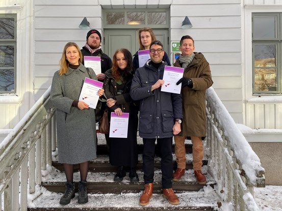 Kulturstipendiaterna står på en trappa utomhus i Härnösand och håller stipendiediplomen framför sig.