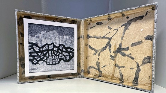 En platt låda, uppslagen som en bok, till vänster i den sitter ett grafiktryck och högra delen är dekorerad med ett mönster. 