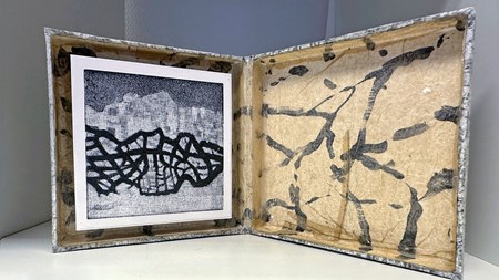 En platt låda, uppslagen som en bok, till vänster i den sitter ett grafiktryck och högra delen är dekorerad med ett mönster. 