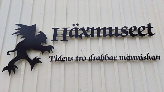 En trävägg med en skylt föreställande häxmuseets logotyp och orden "Häxmuseet, tidens tro drabbar människan"