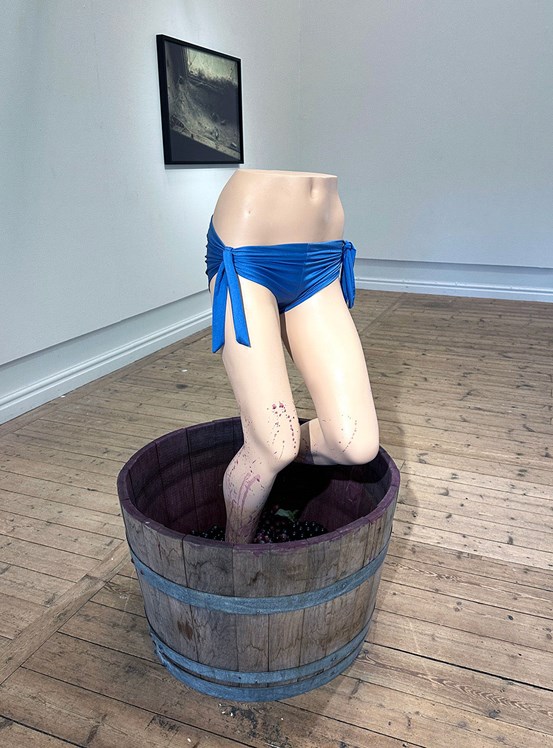 En skyltdocka, i en blå bikini, som saknar överkropp trampar druvor i ett runt träkar. Druvsaft har stänkt på benen. På väggen i bakgrunden syns en bild.