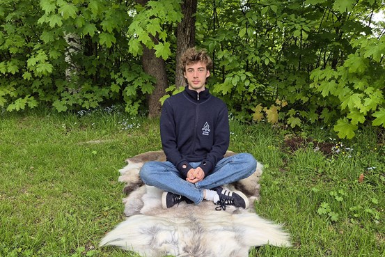 En ung man sitter med korslagda ben på en renfäll ute på en gräsmatta med gröna träd i bakgrunden. 