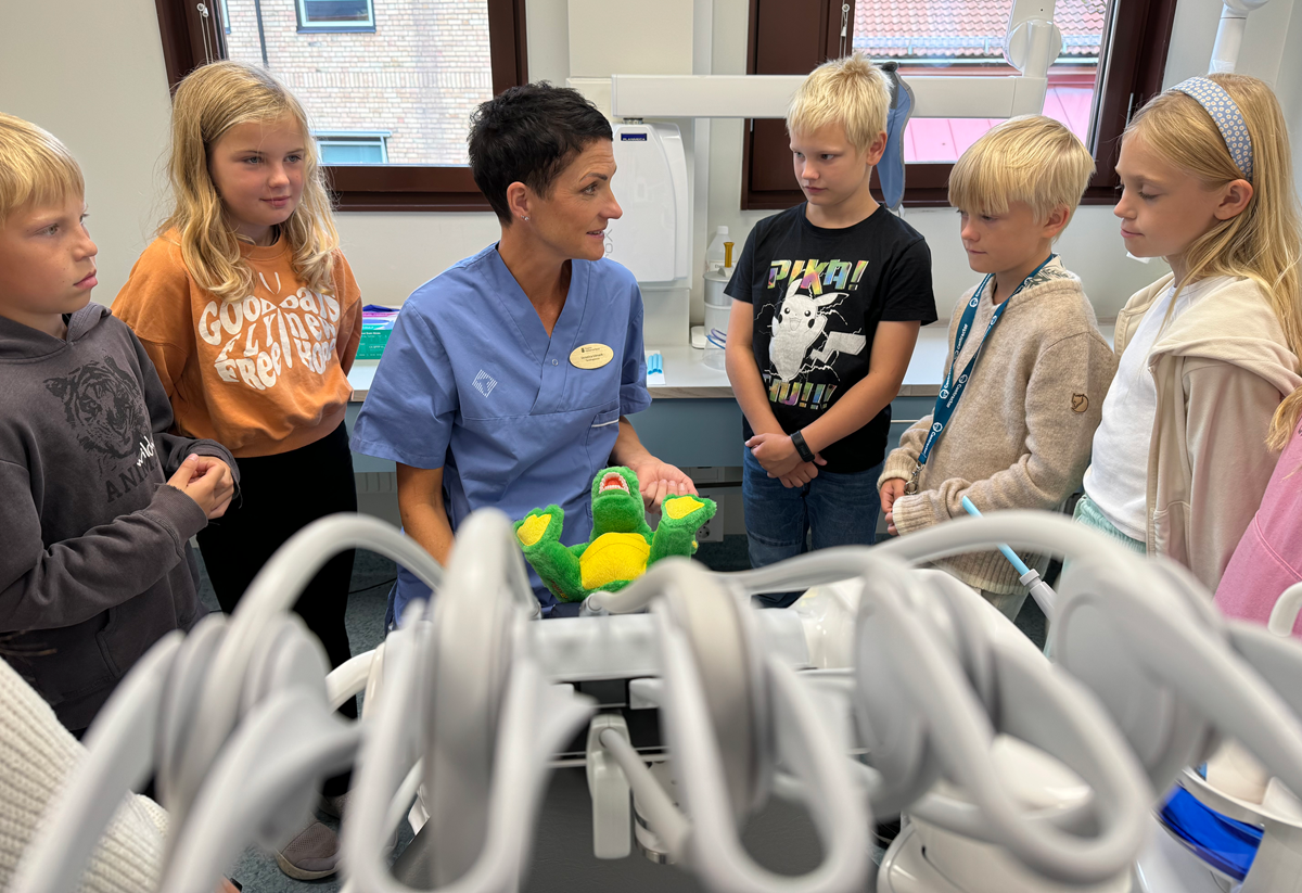 alt='Barn står runt en tandhygienist i ett undersökningsrum.'