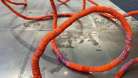 På gråaktiga golvplattor av metall slingrar sig en tjock orange-röd ”kabel” tillverkad av tyg med en röd tråd virad runt för att hålla ihop den. 