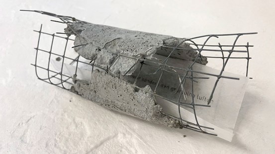 En stål- och betongskulptur formad till en behållare. Metallnät med betong som delvis täcker nätet. Inuti, inkilad, ligger ett papper med text. 