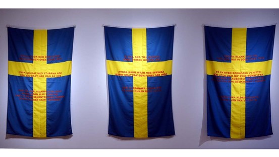 Tre svenska flaggor hänger på rad på en vit vägg. Texter i rött är tryckta på dem.