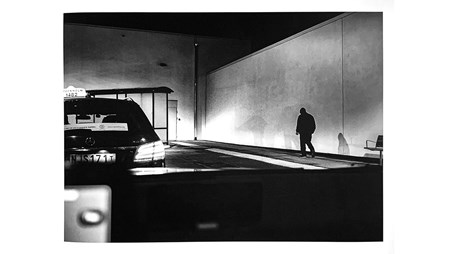 Ett svartvitt foto. Vy från bil på kvällen, genom framrutan. Nertill syns en del av insidan på bilen suddigt. Till vänster framför bilen står en taxi. Till höger en vit vägg, en person går framför väggen. 