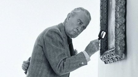 Svartvitt foto av herre med grått hår, mustasch och kostym framåtlutad med förstoringsglas i handen och granskar målning.
