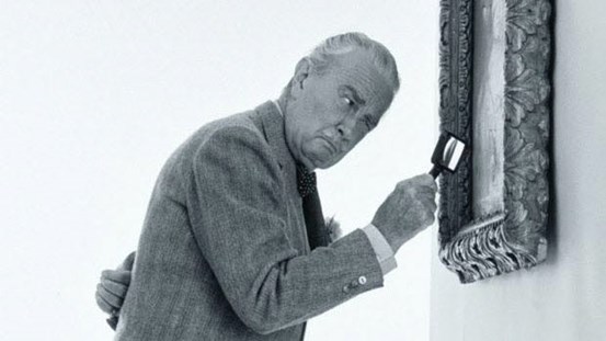 Svartvitt foto av herre med grått hår, mustasch och kostym framåtlutad med förstoringsglas i handen och granskar målning.