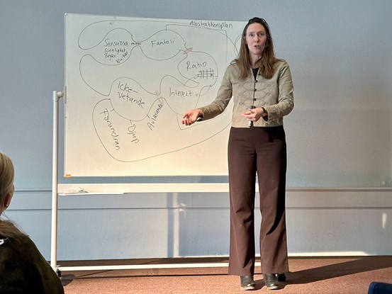 En kvinna med bruna byxor, beige jacka och långt hår står framför en whiteboard. Hon håller en penna i sin vänstra hand och ser ut att prata.