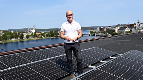 Projektledare vid regionfastigheter Gunnar Bäckman står uppe på taket av Regionens hus en solig dag och vid hans fötter finns nyinstallerade solcellspaneler, man ser utsikten in mot Härnösand och Kyrkviken bakom Gunnar