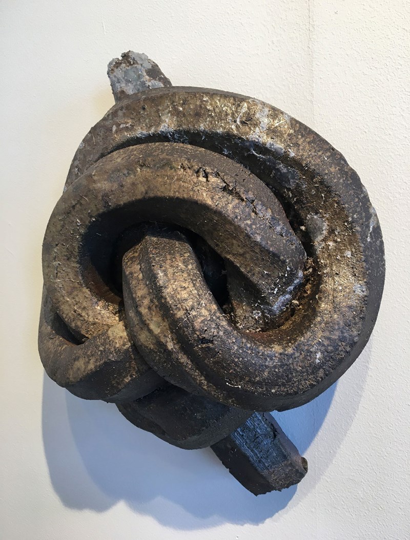 Bilden visar ett vägghängt tredimensionellt keramikverk som representerar en knut. Den mörkbruna knuten är öppen och har ett glänsande skeende som ger den en metallisk aspekt. 