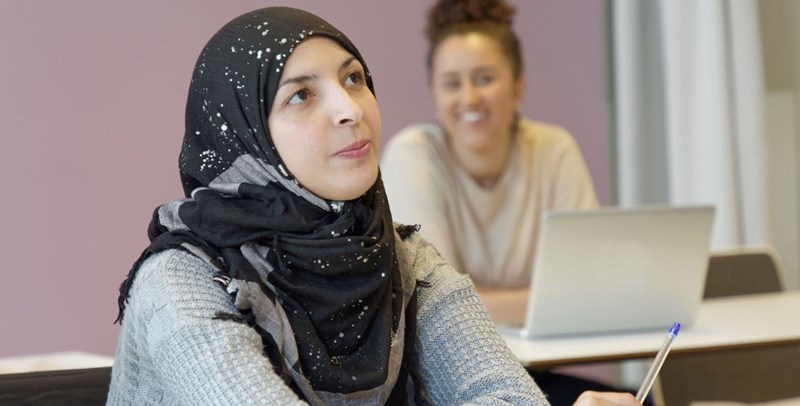 En ung kvinna i slöja tittar drömmande mot taket och en annan ung kvinna sitter bakom med en dator.
