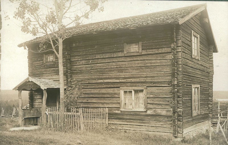 Ett blekt äldre foto i svartvitt. Det föreställer ett torp i timmer. Till vänster en liten farstukvist. Direkt till höger om den är ett litet område med staket runt. Innanför staketet syns ett träd och växter. 