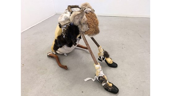Skulptur/objekt liknande en sittande hund. Den är konstruerad av diverse olika material som t.ex.  skumgummi, slitna skor, pinnar, möbelben, pälsstycken, textil, träbitar m.m. 