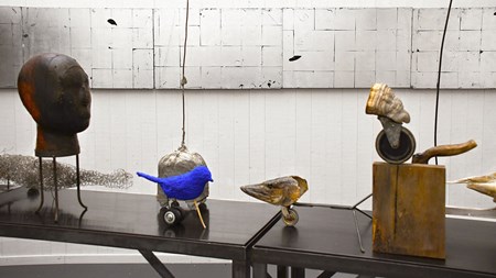 Foto. En serie skulpturala objekt står på några smala avlånga bord. I bakgrunden syns en vit vägg. 