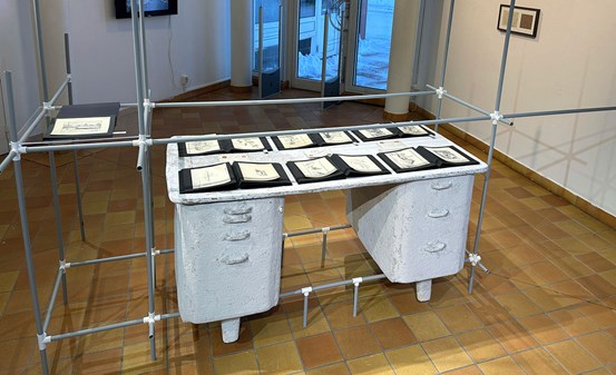 En skrivbord, tillverkat av papier maché, står på en ställningskonstruktion av metallrör. På bordet ligger svarta pärmar uppslagna med pappersark inuti.  