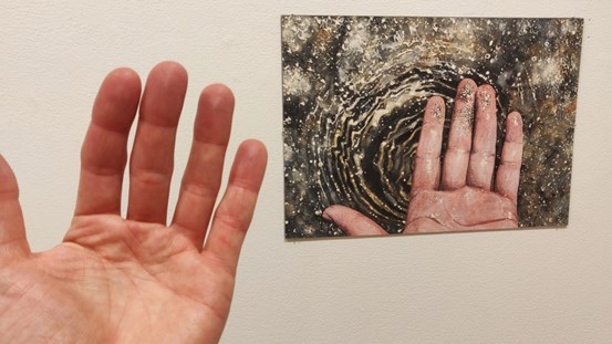 Foto av en hand till vänster om Sara Forsströms målning av en hand som håller i glittrande stoff ovan en vattenyta.