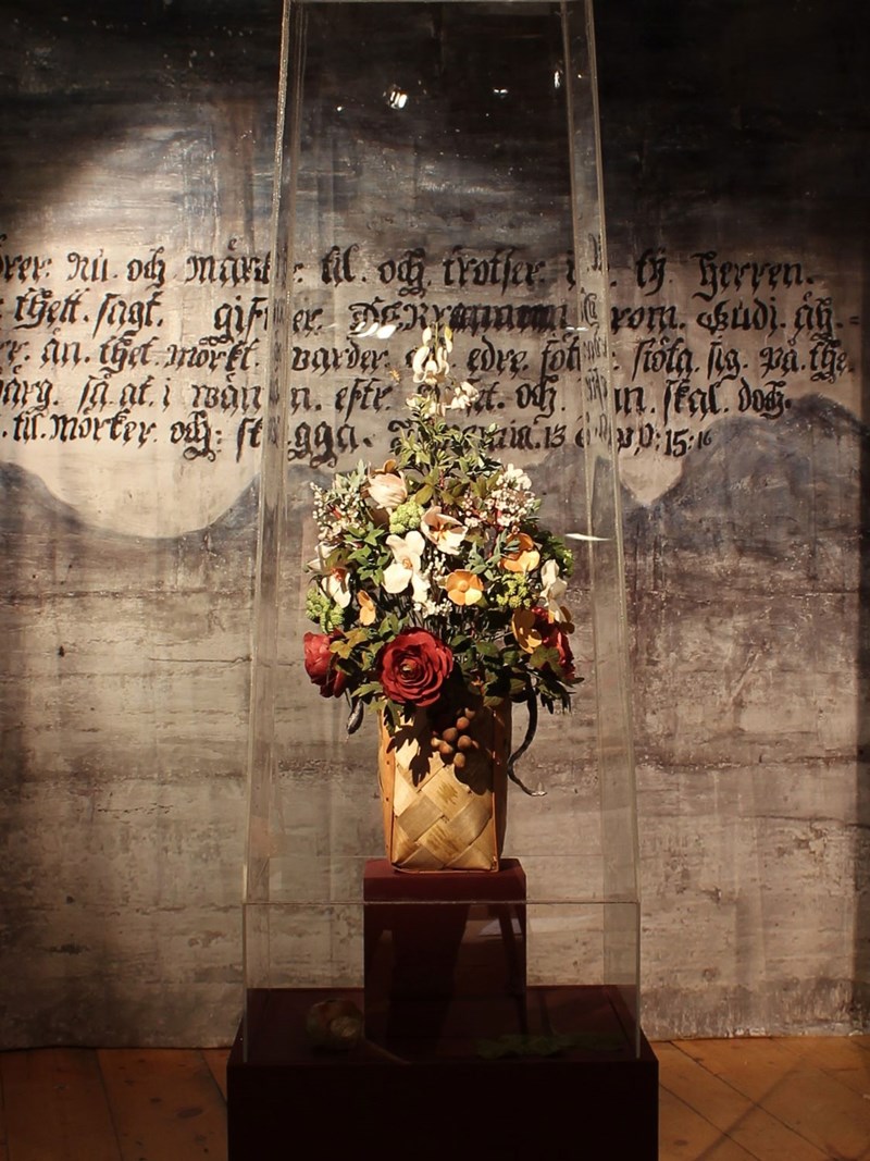 Foto av en stor blomsteruppsättning, gjord av socker, som står på ett podium mitt i bilden. På den grå väggen bakom syns en text skrivet i svart med gammeldags bokstäver. Buketten innehåller blommor i många färger, rött, vitt, gult. 