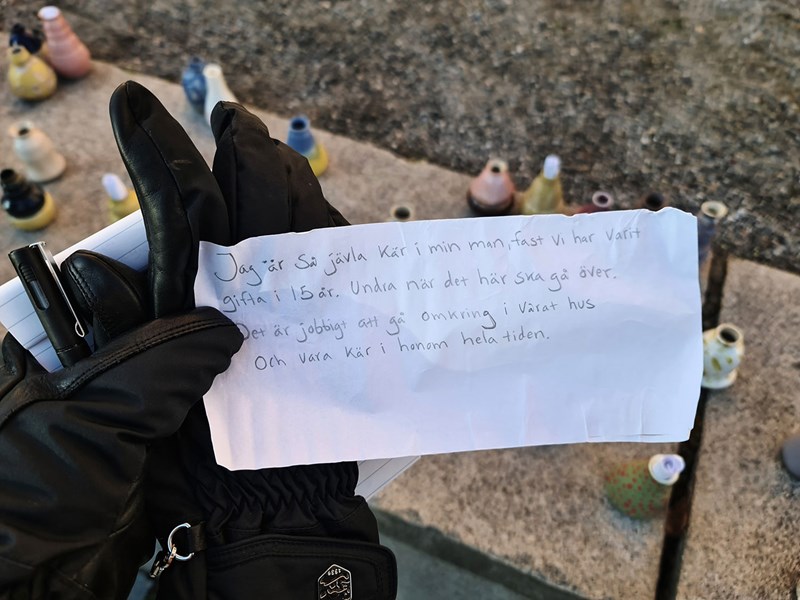 En handskklädd hand håller i en papperlapp med text, en penna och en handske. I bakgrunden står det keramikkärl i olika färger.