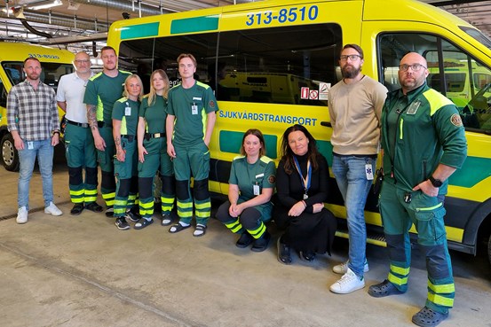 Flera personer, med och utan sjukvårdskläder, står framför en ambulansgul sjuktransportbil.