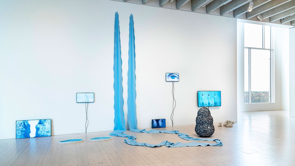 alt='Installation helt i blått, med stenar som svarvats av vatten, en droppformad skulptur klädd med musselskal, ett avlångt tygstycke som sträcker sig mot taket och videoskärmar upphängda på väggen.'