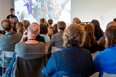 Foto från föreläsning, publik sedd bakifrån framför projektion och föreläsare. 