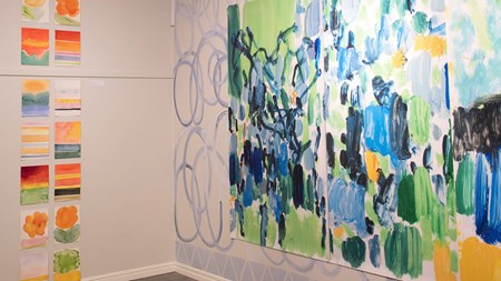 Foto. Vy från utställning. En vägg till höger med en stor färgstark abstrakt målning i mestadels blå och gröna toner men även en del gult. Till vänster syns många mindre målningar i två lodräta rader. 