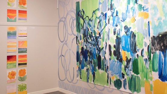 Foto. Vy från utställning. En vägg till höger med en stor färgstark abstrakt målning i mestadels blå och gröna toner men även en del gult. Till vänster syns många mindre målningar i två lodräta rader. 