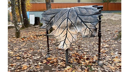 Gulbruna löv ligger på marken runt en skulptur, ett stort lönnlöv liggandes över en ställning (båda i metall). 