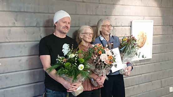 Tre personer som håller i blombuketter framför en grå vägg. 