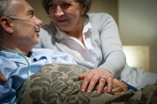 Äldre par som håller i varandras händer och ser på varandra, mannen ligger i en sjukhussäng och en kvinna finns vid hans sida