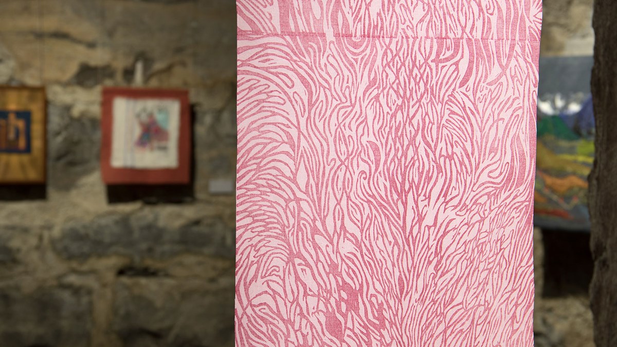 alt='I förgrunden ett träsnitt på tyg föreställande rosa organiska former tryckta på ett vitt tyg. I bakgrunden skymtar en grå stenvägg med fler verk uppsatta. '