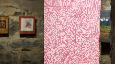 I förgrunden ett träsnitt på tyg föreställande rosa organiska former tryckta på ett vitt tyg. I bakgrunden skymtar en grå stenvägg med fler verk uppsatta. 
